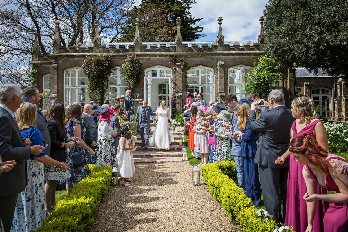 Newlyweds walk hand-in-hand down a gravel path outside a stone orangery as guests shower them with confetti during a summer wedding.