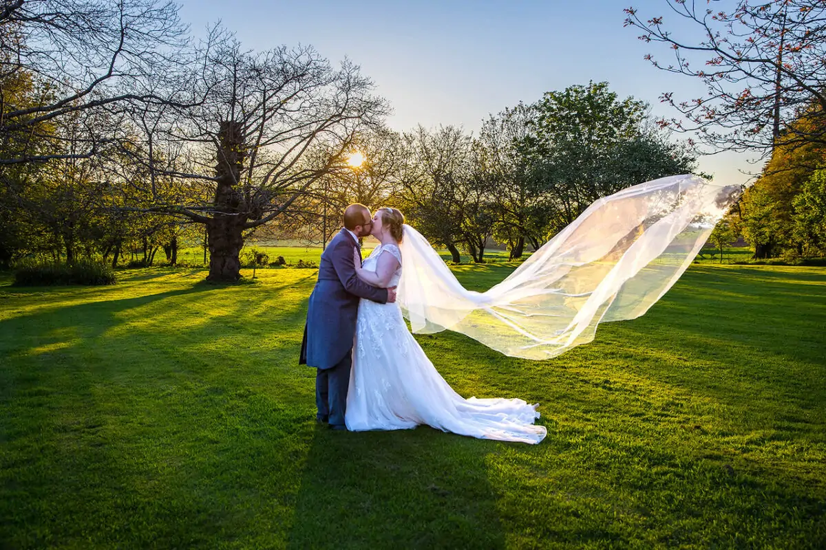 A newlywed couple shares a tender kiss in a lush green park during golden hour, with the bride's long cathedral veil dramatically catching the breeze.