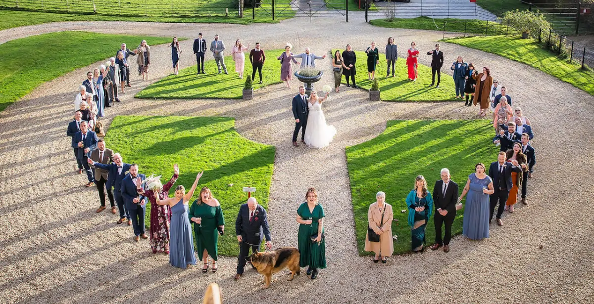 An elevated drone shot captures the bride and groom standing in the centre of a large gravel driveway flanked by manicured lawns, surrounded by a large group of cheering wedding guests and a German Shepherd.