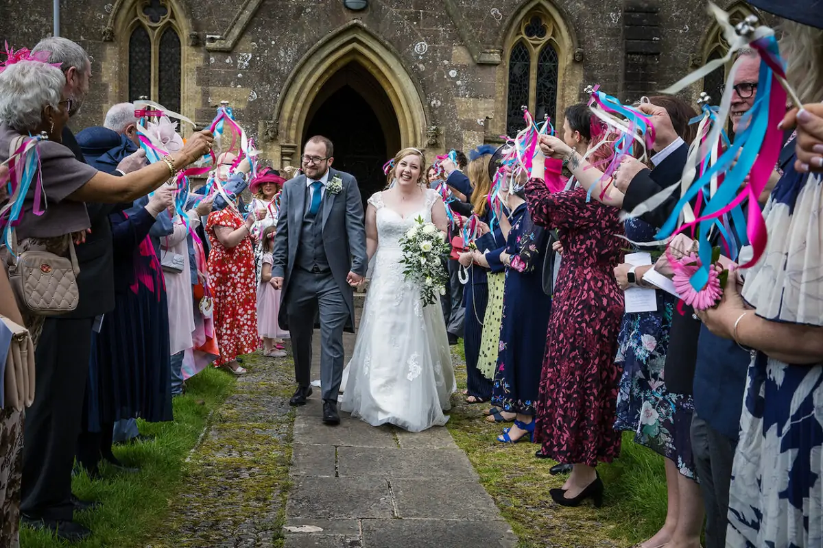 A newlywed couple smiling broadly as they walk out of a stone church hand in hand, cheered on by wedding guests waving colourful ribbon wands.