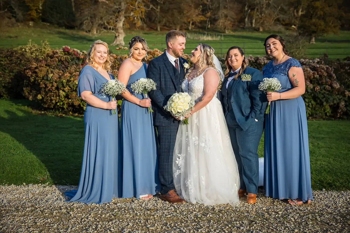 A smiling bridal party in dusty blue dresses and a tweed suit poses outdoors in a sunlit English country garden.