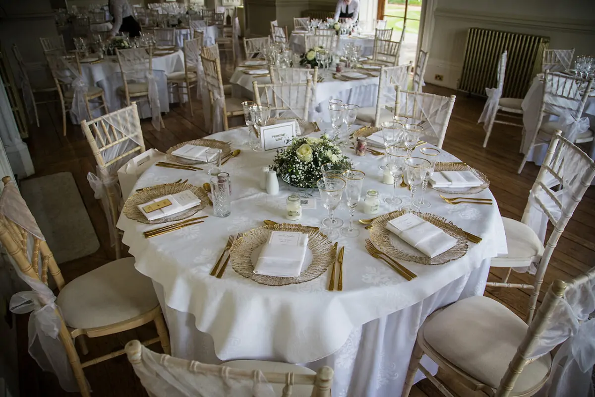 An elegantly set round wedding reception table features crisp white linen, gold charger plates, crystal glassware, and a delicate floral centrepiece surrounded by Chiavari chairs.