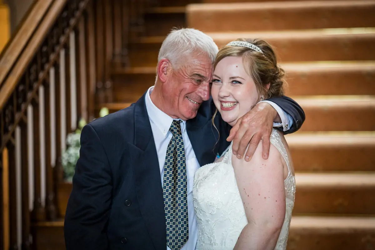 A smiling bride shares an emotional, tender moment with her father on a grand carpeted staircase inside a venue.