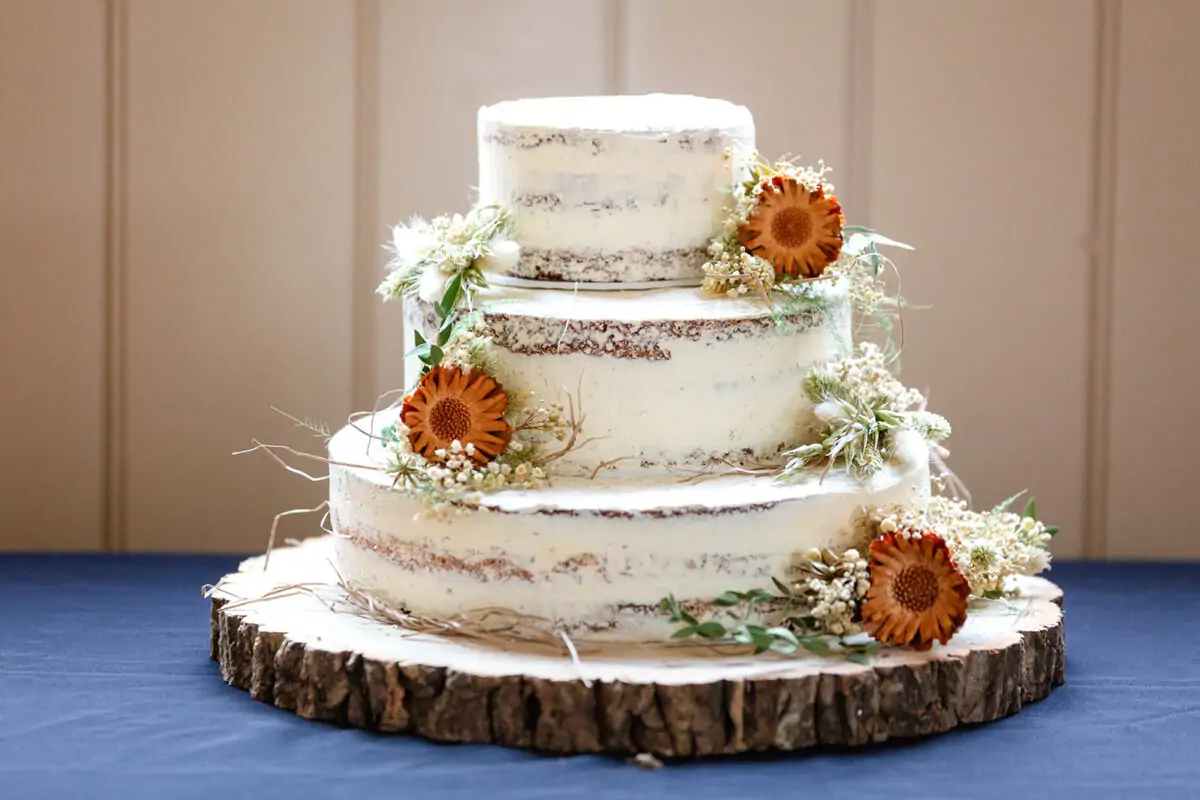 A three-tier semi-naked wedding cake decorated with dried autumn flowers and foliage sits upon a rustic wood slice stand.