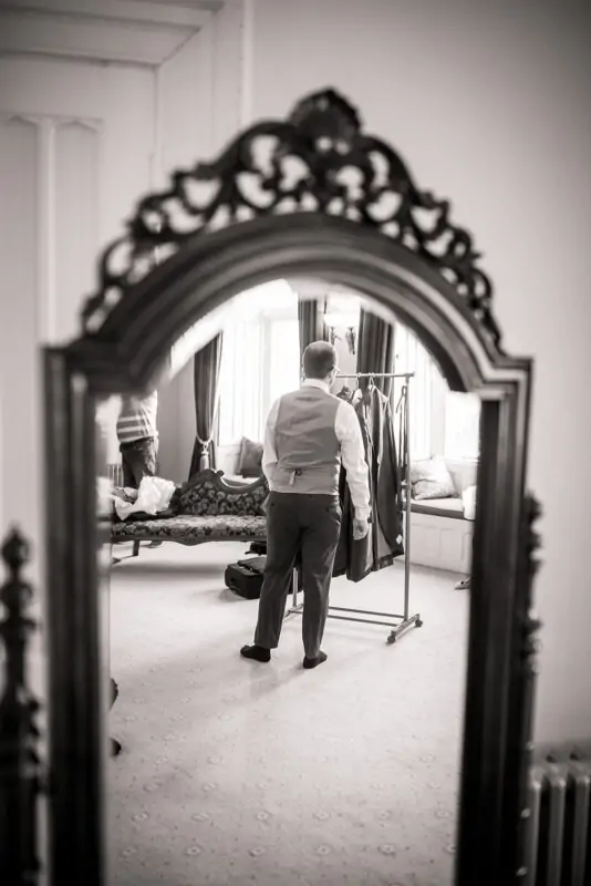 A groom seen from behind in a mirror reflection as he prepares in a light-filled room during morning wedding preparations.