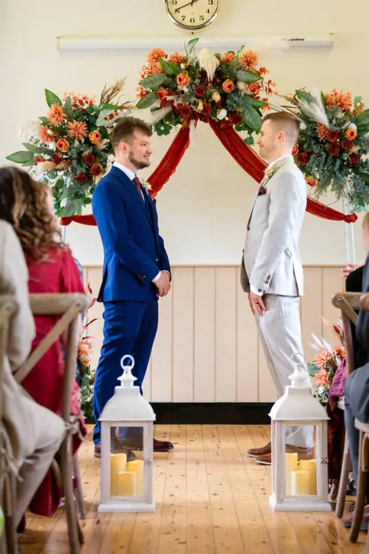 Two grooms stand facing each other during a civil ceremony in front of a rustic floral arch adorned with autumn-toned flowers and white lanterns lining the aisle.