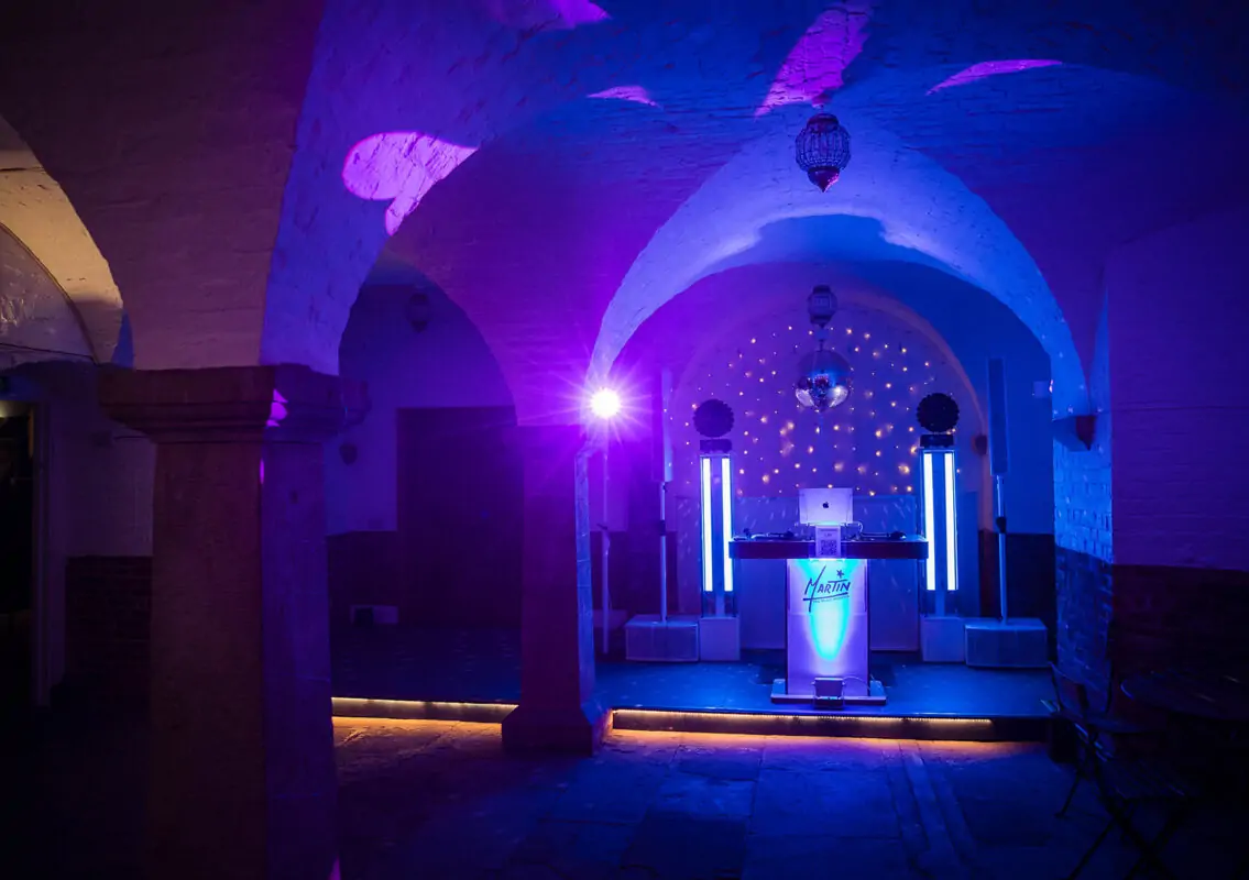 An empty vaulted brick venue illuminated with dramatic blue and purple stage lighting featuring a DJ booth and disco ball.