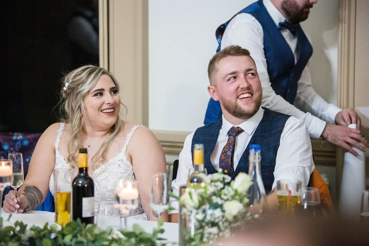 A newlywed couple laughing joyfully at the top table during wedding speeches, surrounded by wine bottles, candles and floral table arrangements.