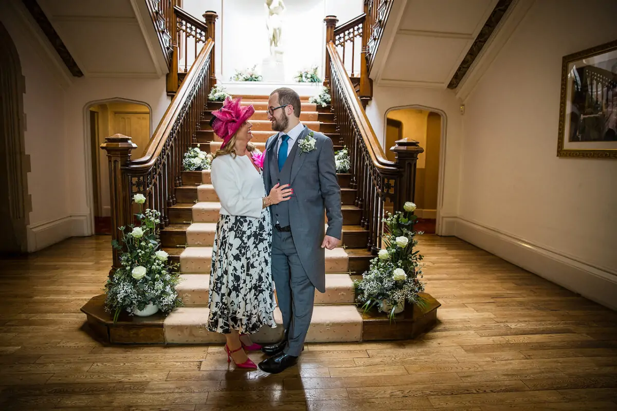 An older couple poses affectionately at the base of a grand wooden staircase inside a magnificent manor house venue.