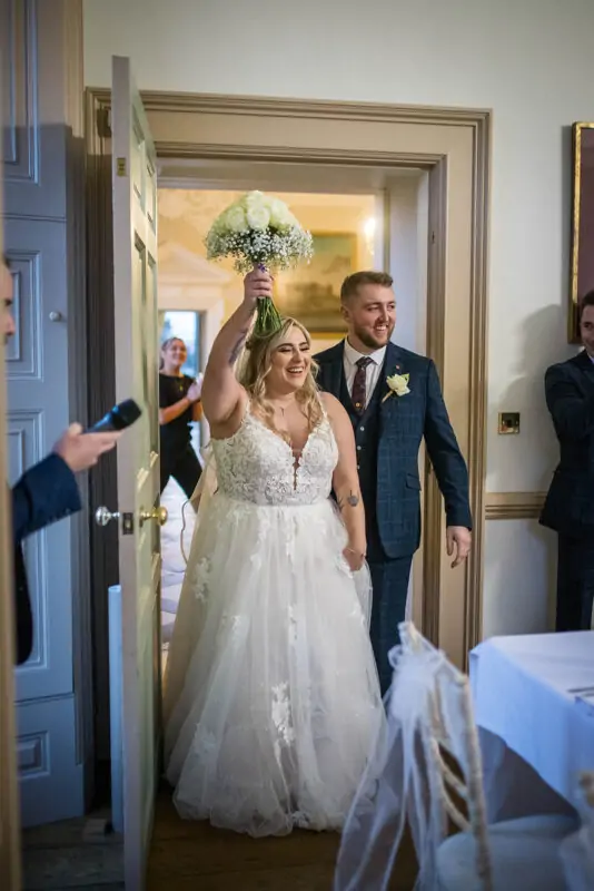 The joyful bride and groom make their grand entrance into the reception room, with the laughing bride raising her white rose bouquet triumphantly in the air.