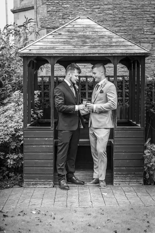 Two grooms stand inside a wooden garden gazebo in smart suits, clinking glasses of champagne together on their wedding day.