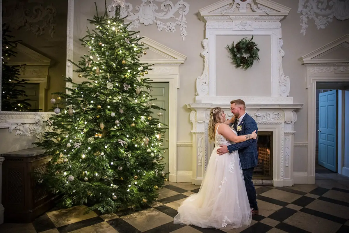 A bride and groom share a tender forehead kiss in a grand country house decorated for a winter wedding, standing next to a large illuminated Christmas tree and a traditional fireplace.