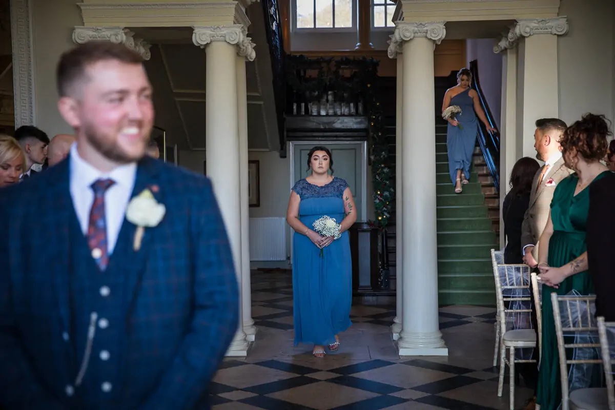 A smiling groom in a blue checked suit waits at the front of a grand manor house hall while bridesmaids walk down a sweeping staircase in dusty blue dresses.