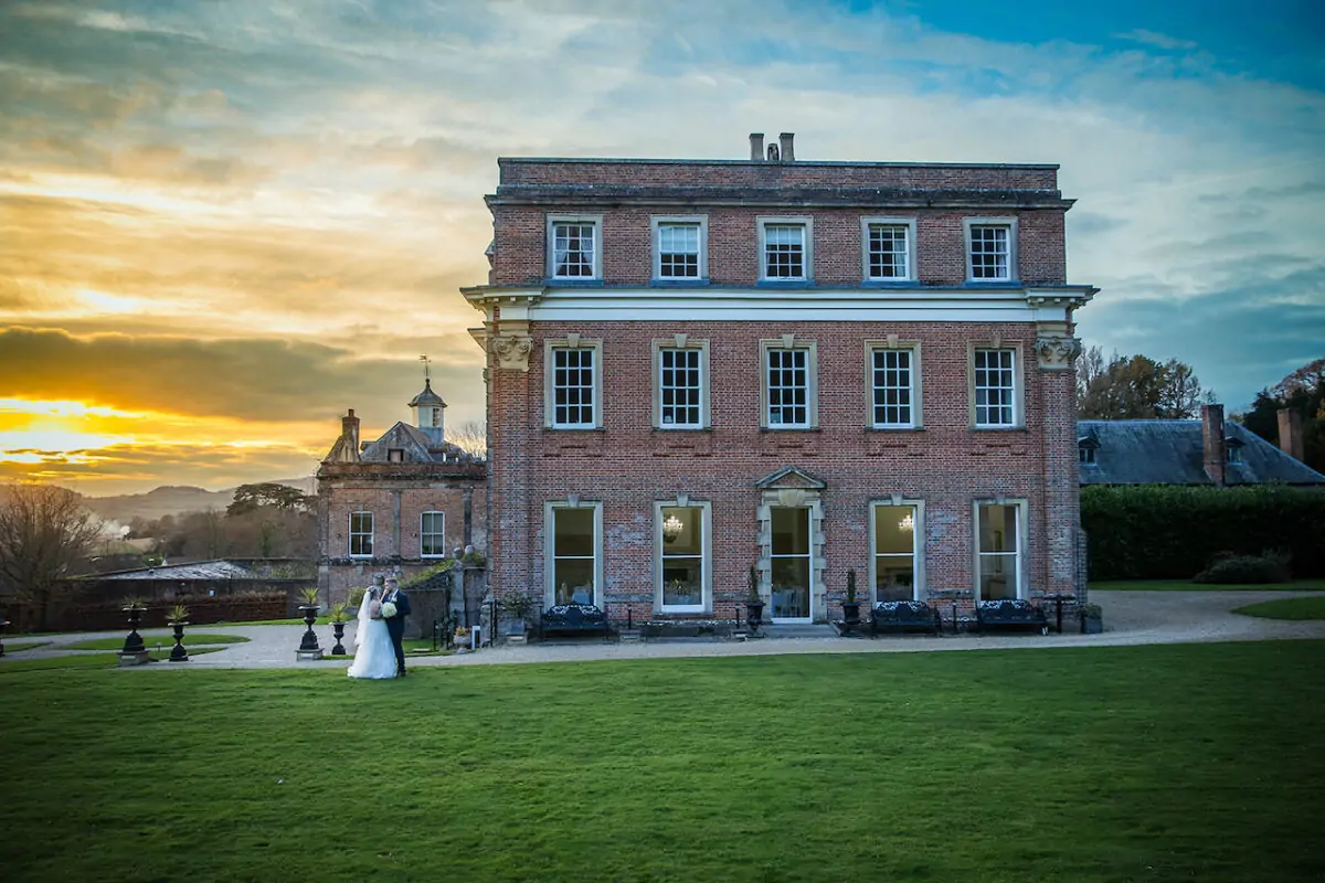 A newlywed couple stands on a sprawling lawn before a grand brick country house during a breathtaking golden sunset.