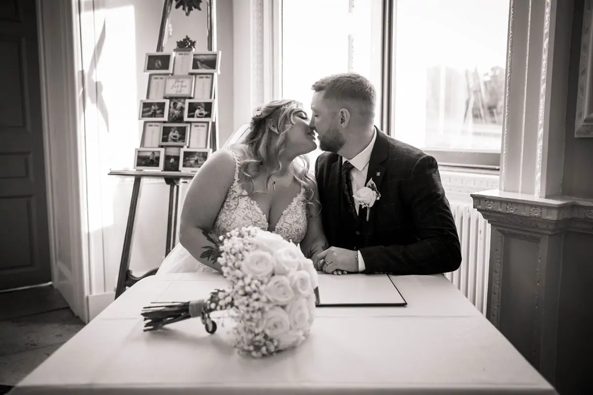 A newlywed couple share a tender kiss over the signing table next to a floral bouquet and a photo seating plan.