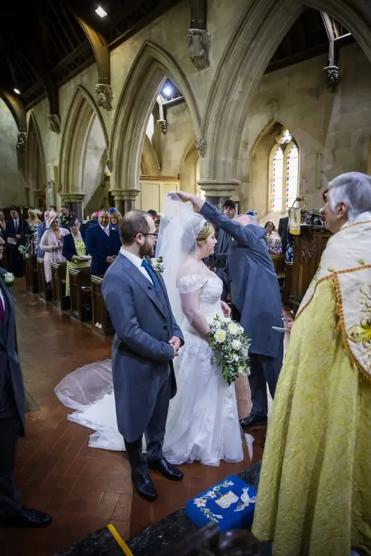A bride in an off-the-shoulder a-line gown and a groom in a morning suit stand at the altar during a traditional church ceremony.