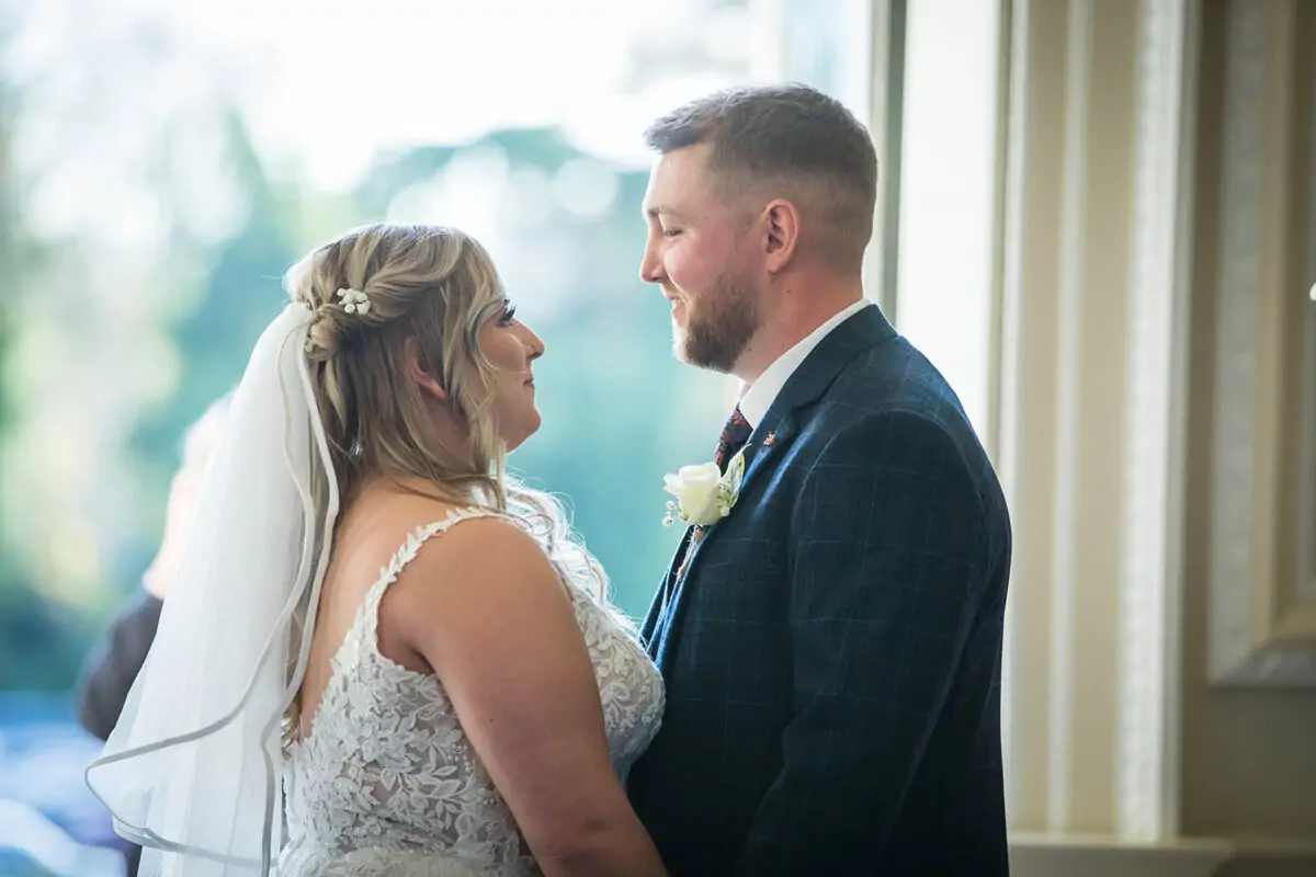 A newlywed couple gazes lovingly at each other indoors near a bright window, with the bride wearing a lace gown and veil.
