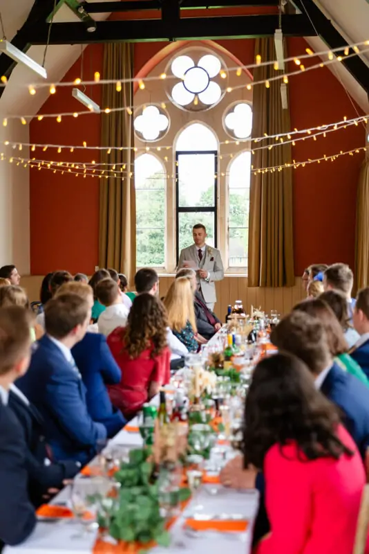 A groom delivers a speech standing at the end of a long banquet table decorated with foliage inside a village hall with vaulted timber ceilings and tall windows.