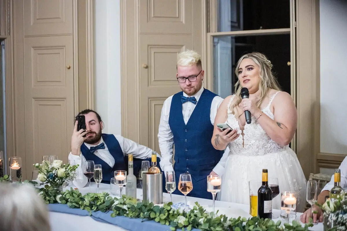 A smiling bride delivers her speech at the top table, holding a microphone while standing next to a groomsman in a blue waistcoat inside a wood-panelled room.