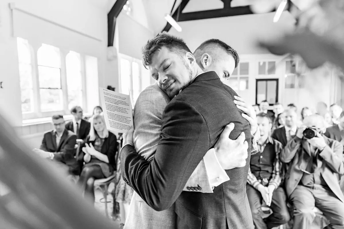 A black and white photograph captures two grooms sharing an emotional embrace during their wedding ceremony as one holds a letter.