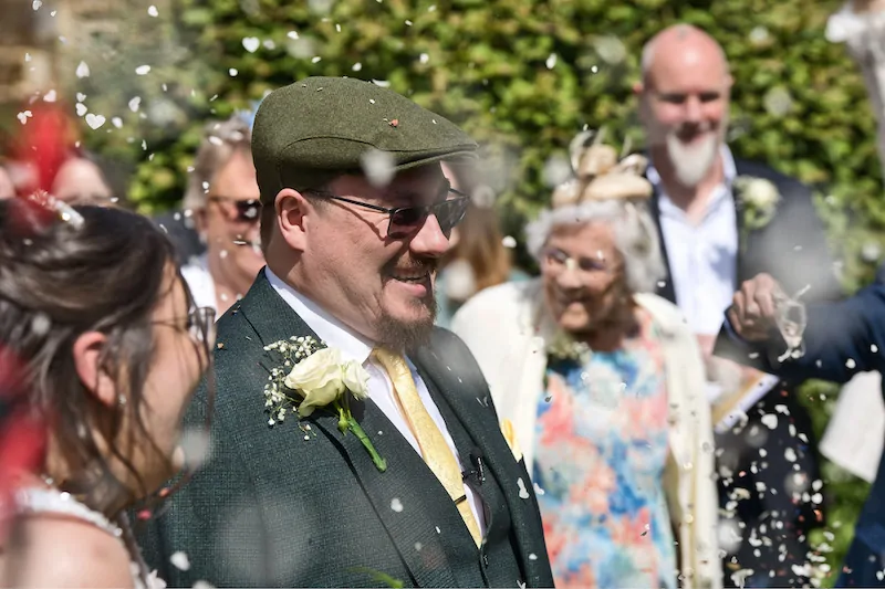 A smiling groom wearing a green tweed suit and flat cap laughs as guests shower the newlyweds with a flurry of white confetti.