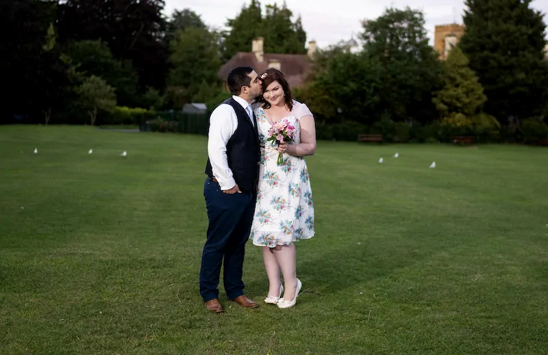 A groom gently kisses the bride's forehead in a lush green garden while she holds a colourful floral bouquet.