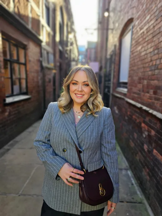 A smiling guest with blonde wavy hair poses in a grey pinstripe blazer while holding a dark shoulder bag in a narrow brick alleyway.