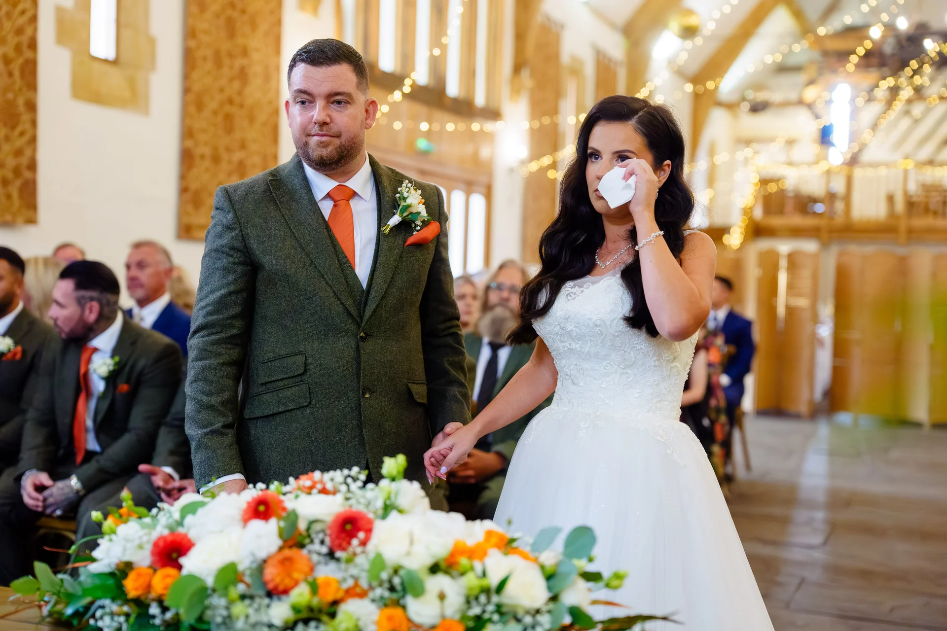 An emotional bride wipes away happy tears with a tissue during the ceremony while holding hands with her groom at the altar.