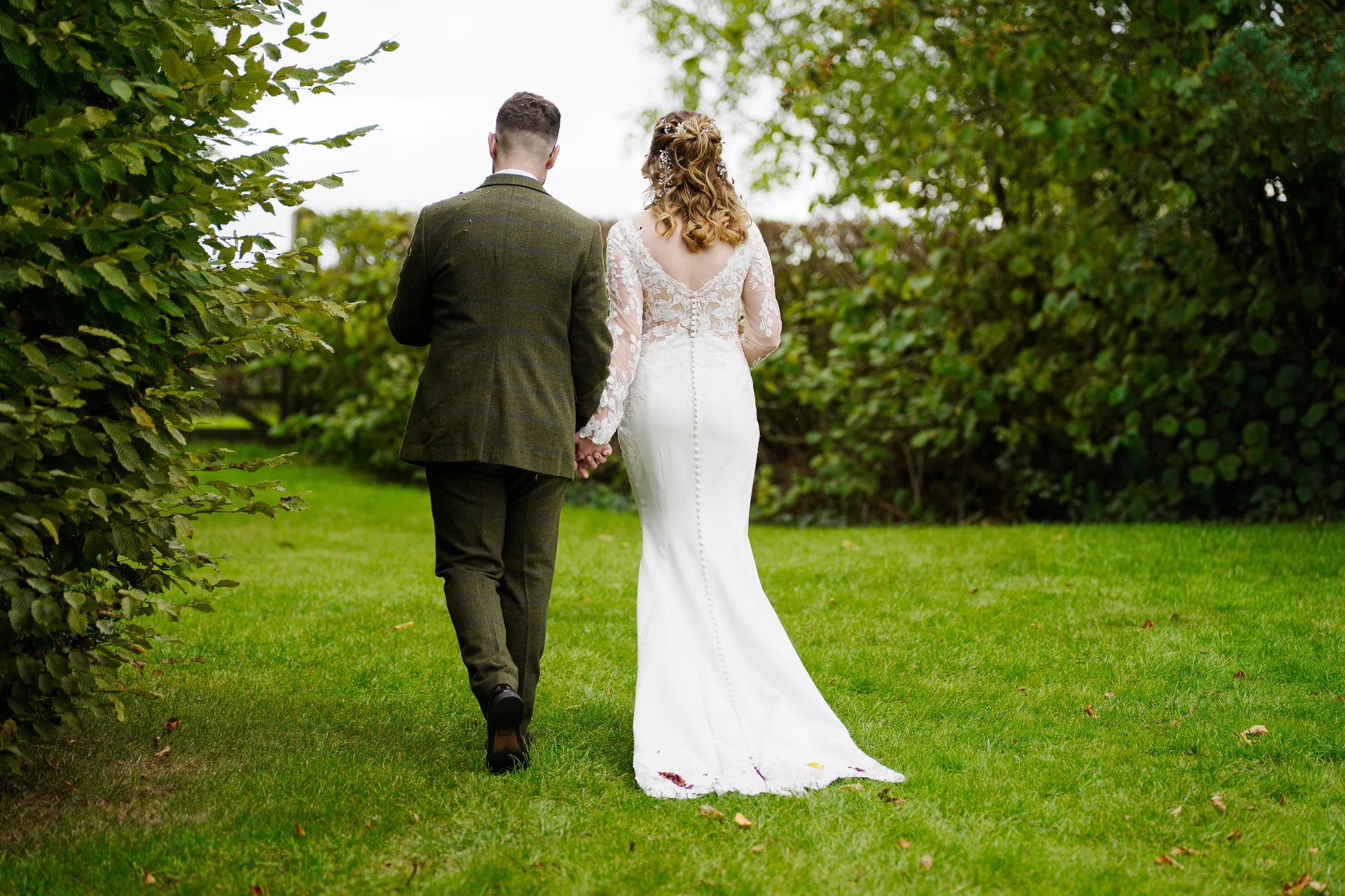 A newlywed couple is captured from behind walking hand in hand across a lush green lawn surrounded by trees wearing a tweed suit and a fitted lace-detailed wedding dress.