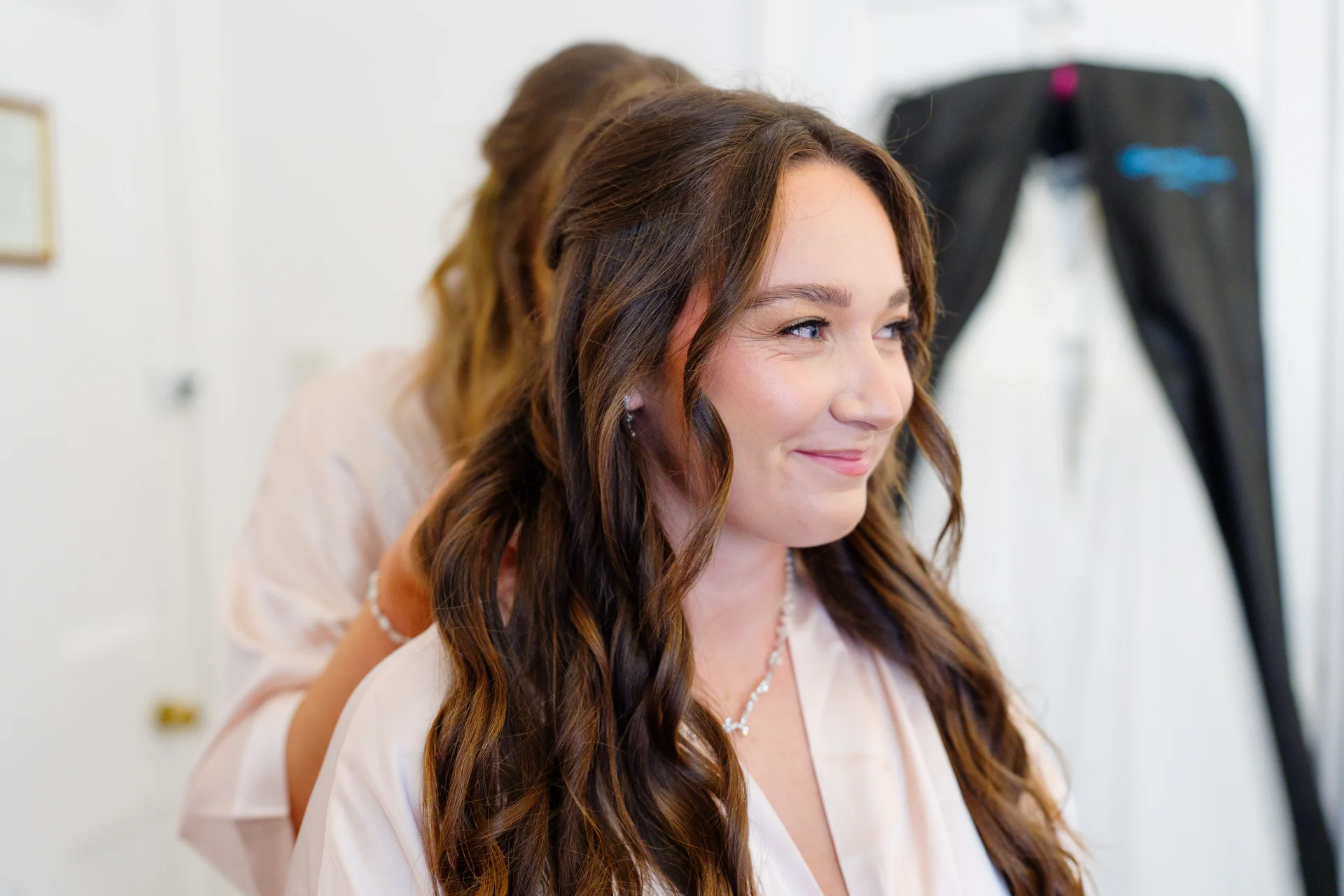 A smiling bride with long, wavy brown hair gets her hair styled during morning bridal preparations while wearing a silky robe.