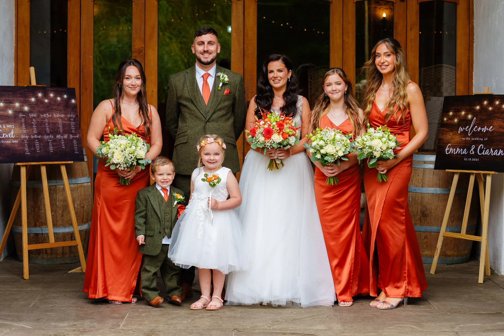 A bridal party poses outdoors in front of wooden doors, with the bride in a white gown holding a vibrant bouquet, flanked by bridesmaids in rust satin dresses, a groom in a green tweed suit, and young children in matching attire.
