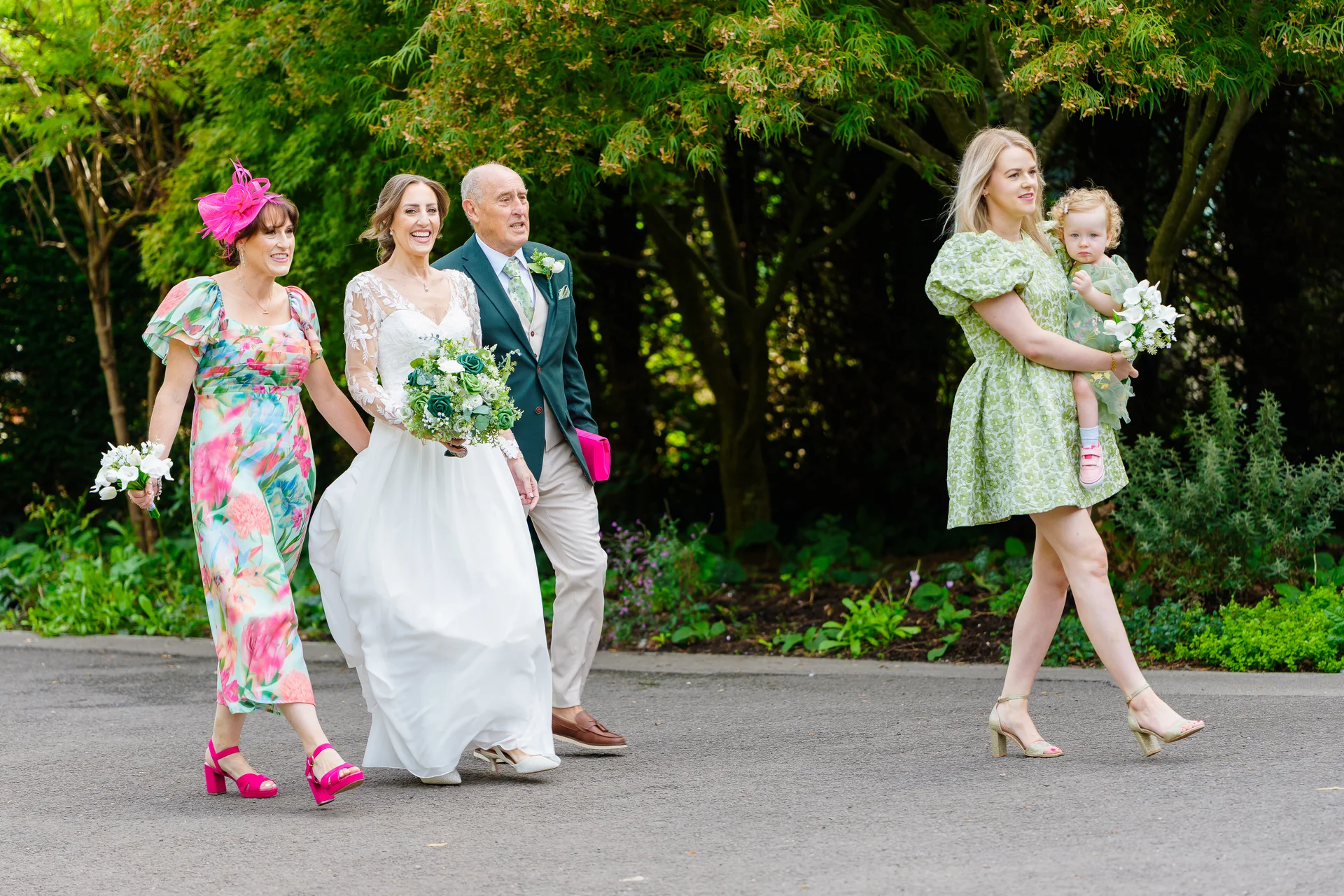 A smiling bride walking outdoors with her father and mother alongside other guests and a young flower girl holding a small bouquet.