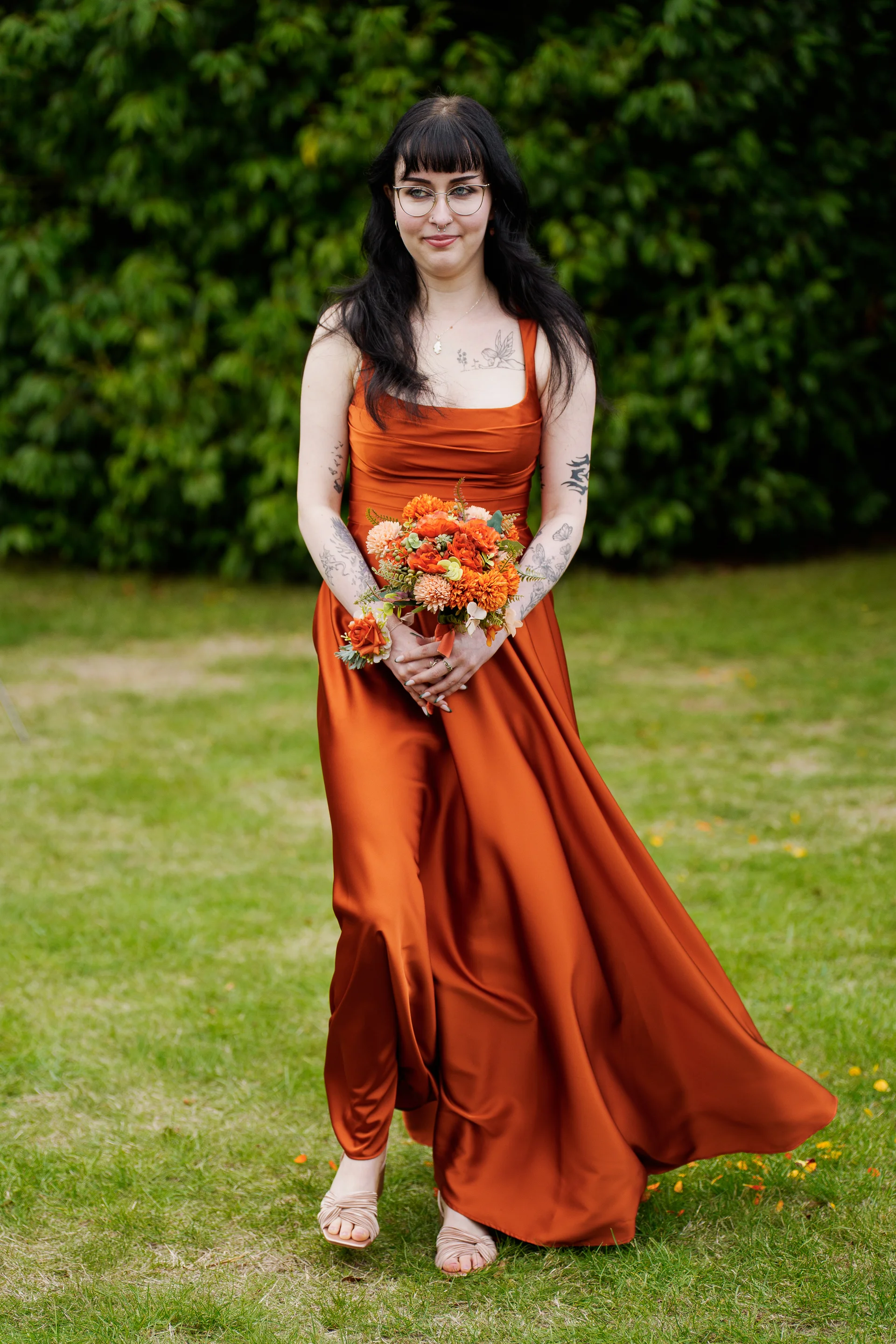 A solo bride with long dark hair and tattoos poses on a green lawn wearing an elegant rust-orange satin gown and holding a vibrant autumn-toned bouquet.