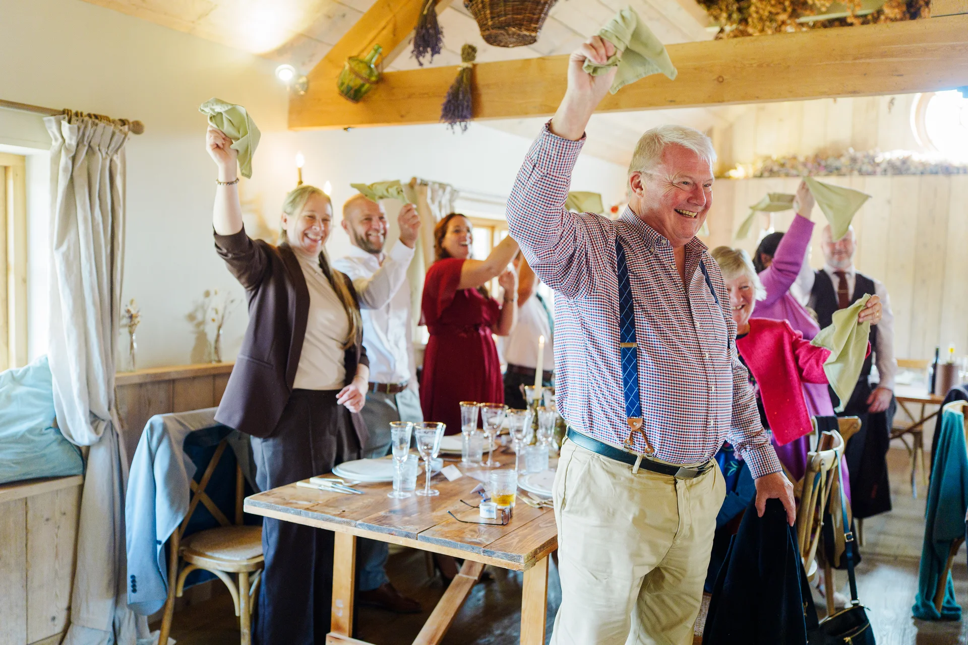 Wedding guests joyfully wave their napkins in the air around a wooden banquet table inside a rustic barn reception venue.