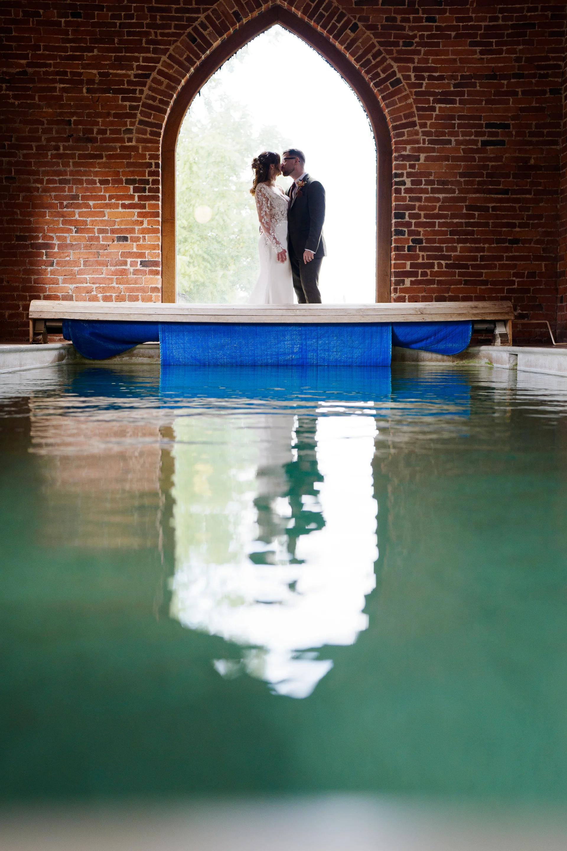 A bride and groom share a tender kiss framed inside an arched brick window, with their reflection shimmering across the calm water below.