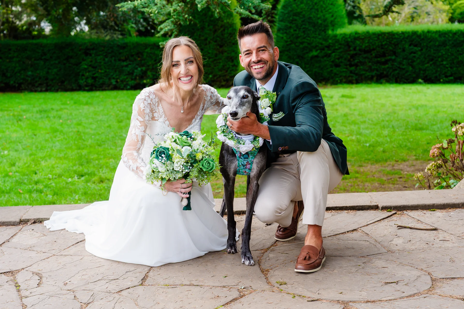 A smiling bride in a long-sleeved lace wedding dress and a groom in a green suit pose happily outdoors with their greyhound wearing a floral collar.