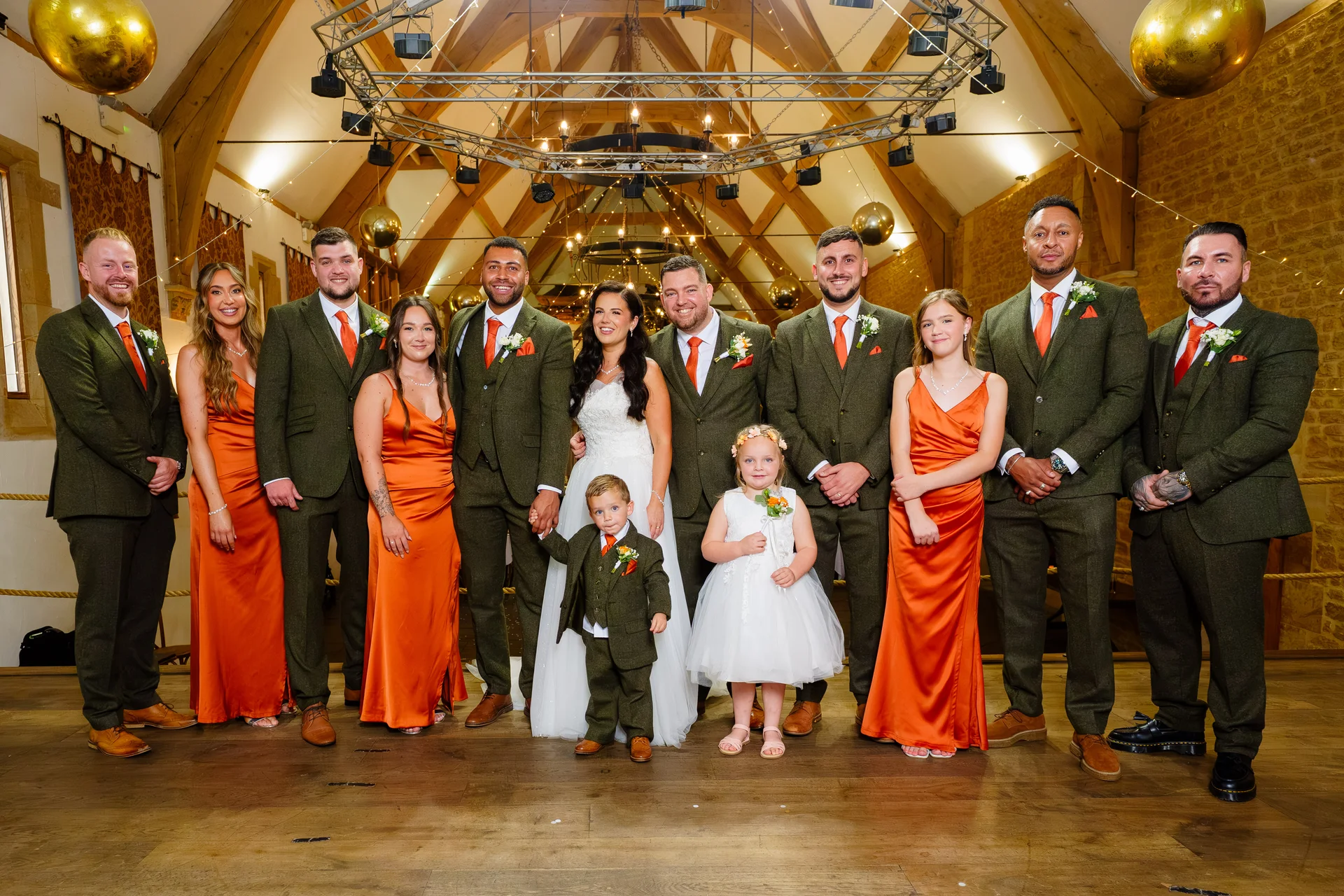 A large wedding party including the bride and groom, groomsmen in green tweed suits with orange ties, bridesmaids in orange silk slip dresses, and two young children posing on a wooden stage inside a rustic stone barn.