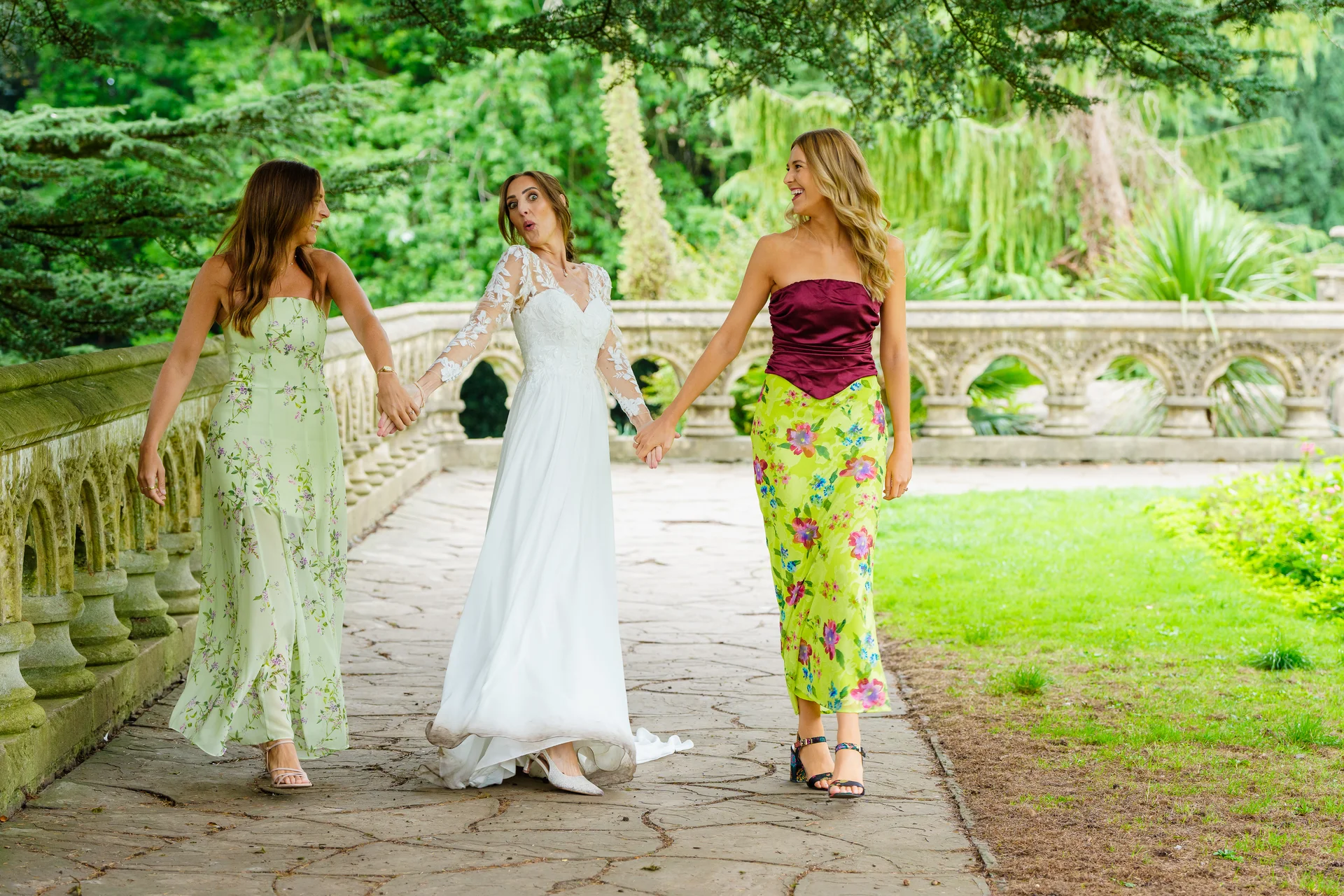 A laughing bride in a long-sleeved white gown walks hand-in-hand along a stone bridge with two bridesmaids wearing colourful floral dresses.