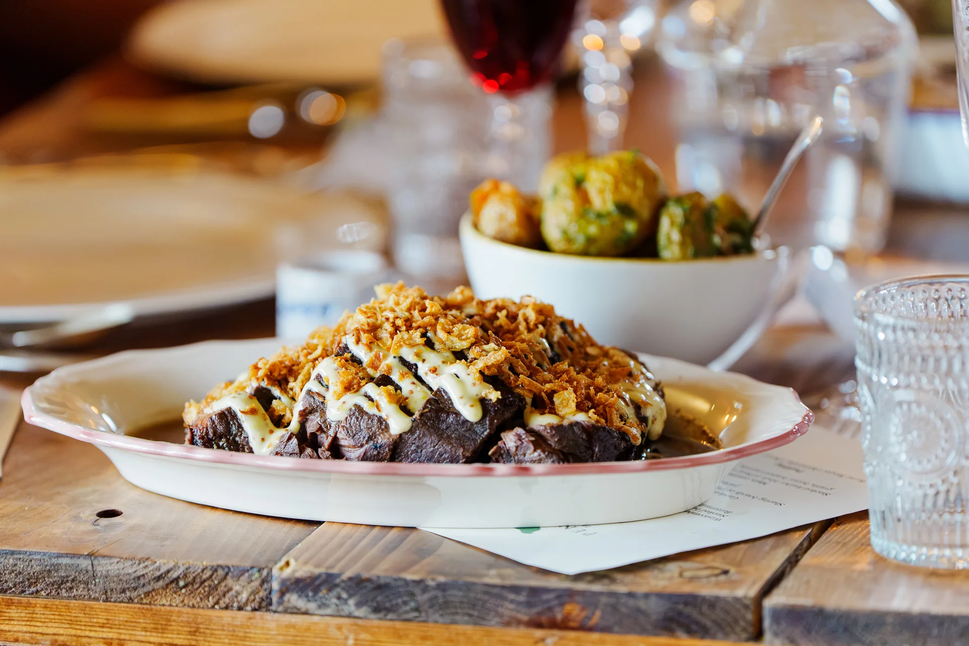 A rustic sharing platter of slow-cooked meat topped with crispy onions and a rich sauce sits on a wooden banquet table at a wedding reception.