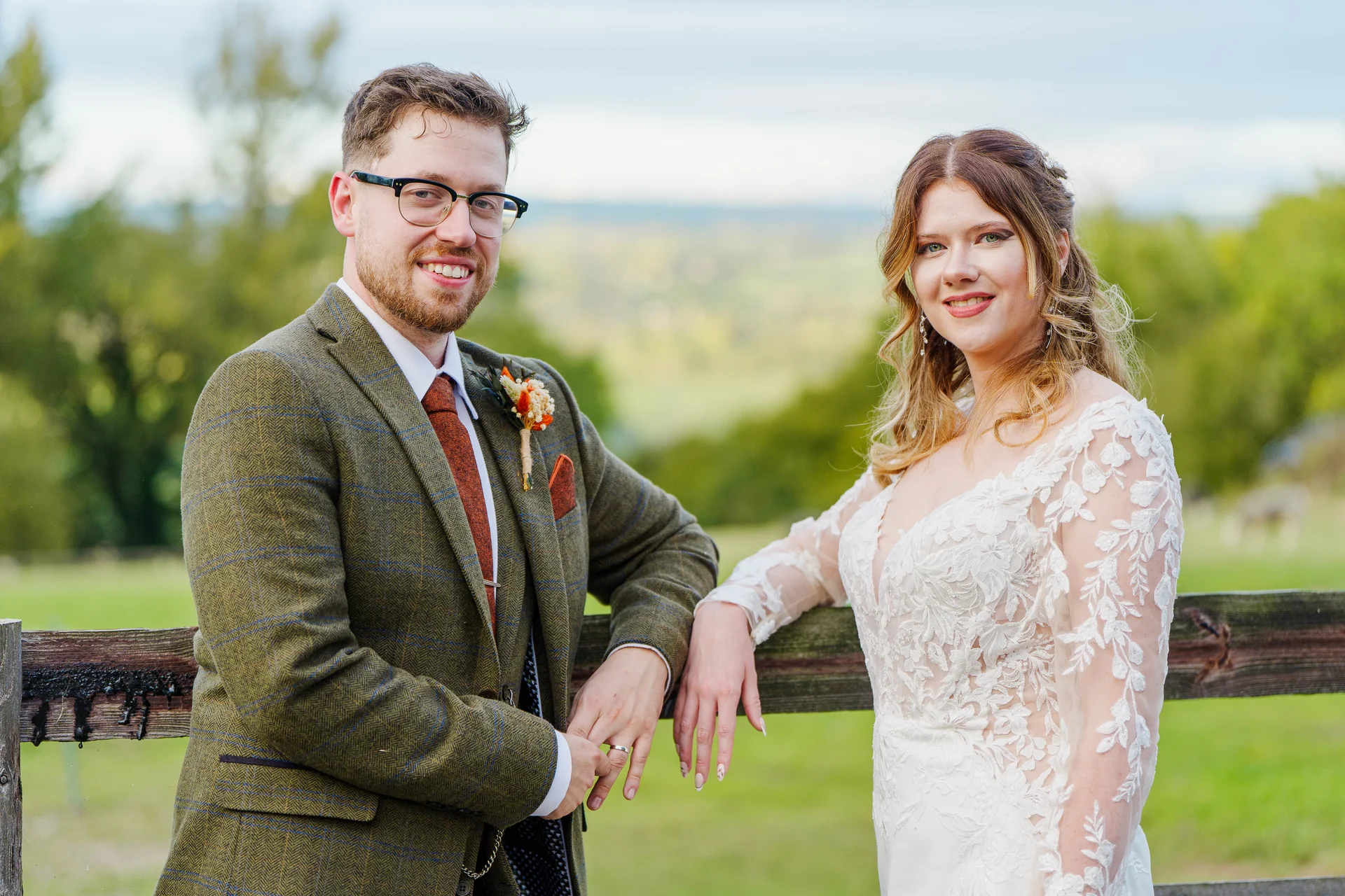 A smiling bride and groom pose together outdoors, leaning against a wooden fence with rolling green hills in the background.