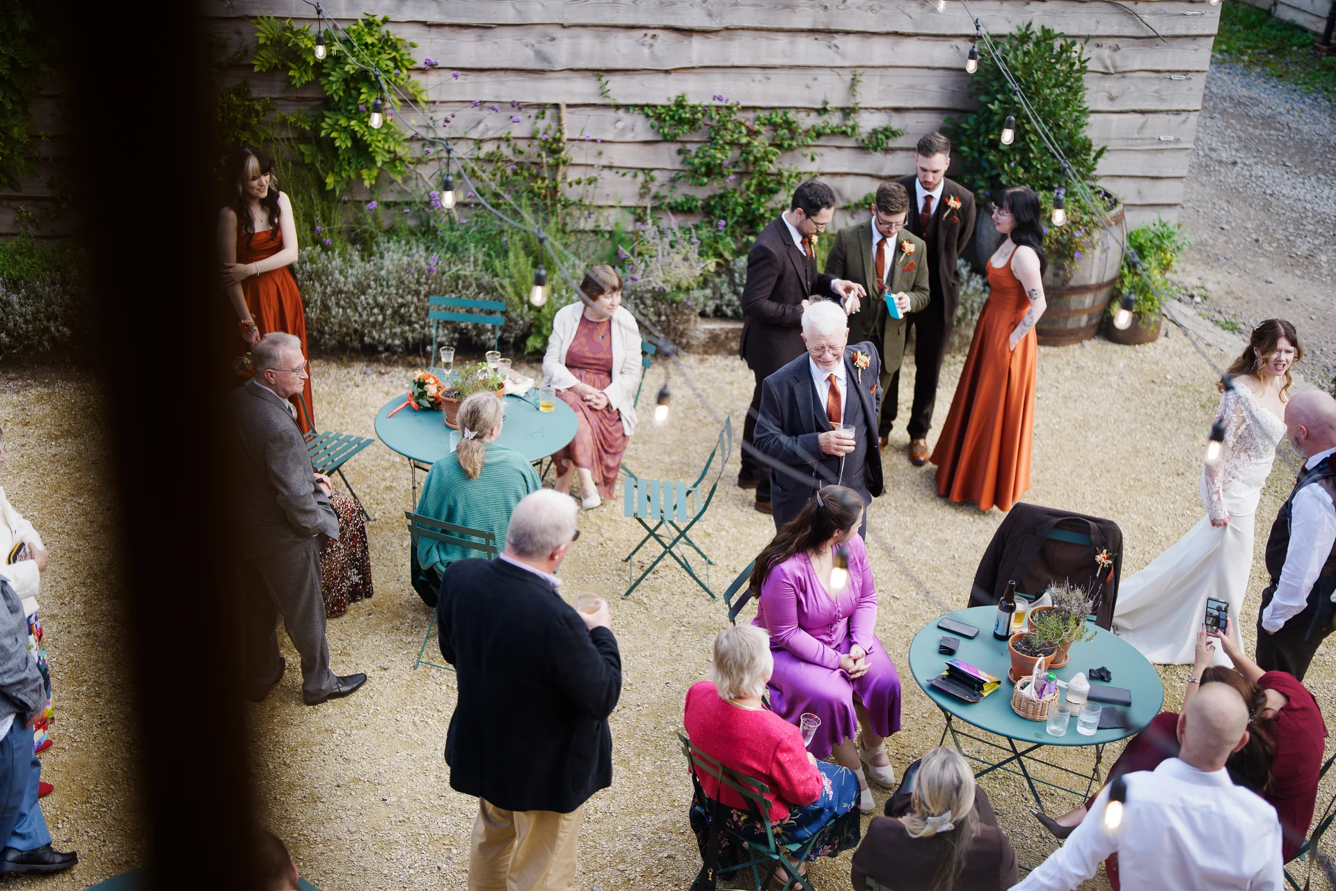 An elevated view of wedding guests mingling in an outdoor gravel courtyard illuminated by festoon lights during the reception.