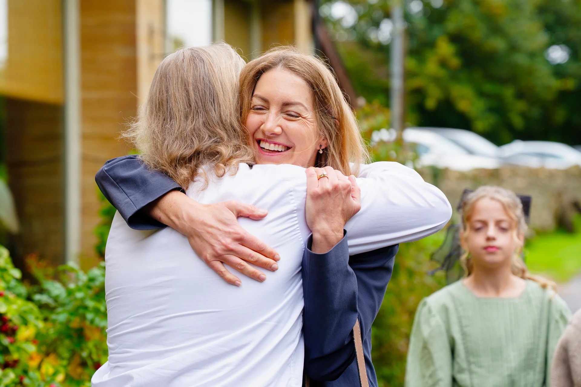 A smiling woman shares a warm, joyful embrace with another person outdoors during a wedding celebration.