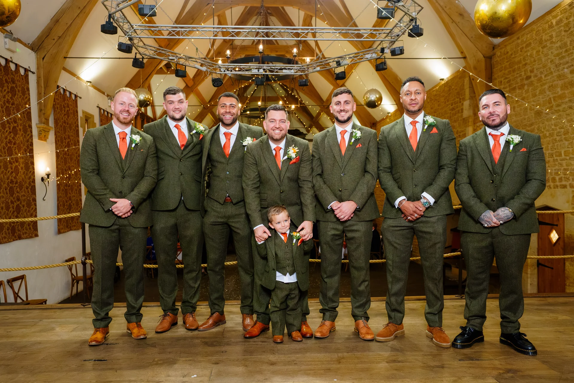 A groom and his groomsmen stand proudly together in matching olive green tweed suits with orange ties inside a rustic barn venue with exposed wooden beams.