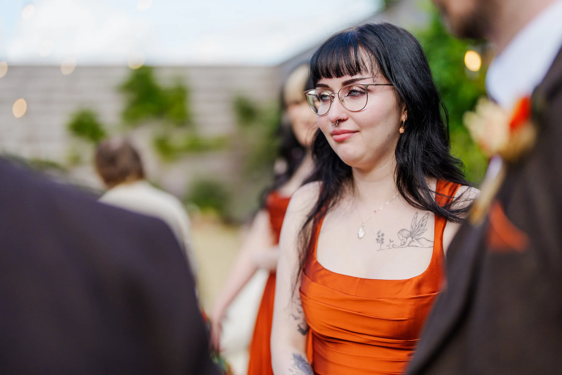 A guest wearing glasses and a burnt orange dress looks on emotionally during an outdoor wedding reception.
