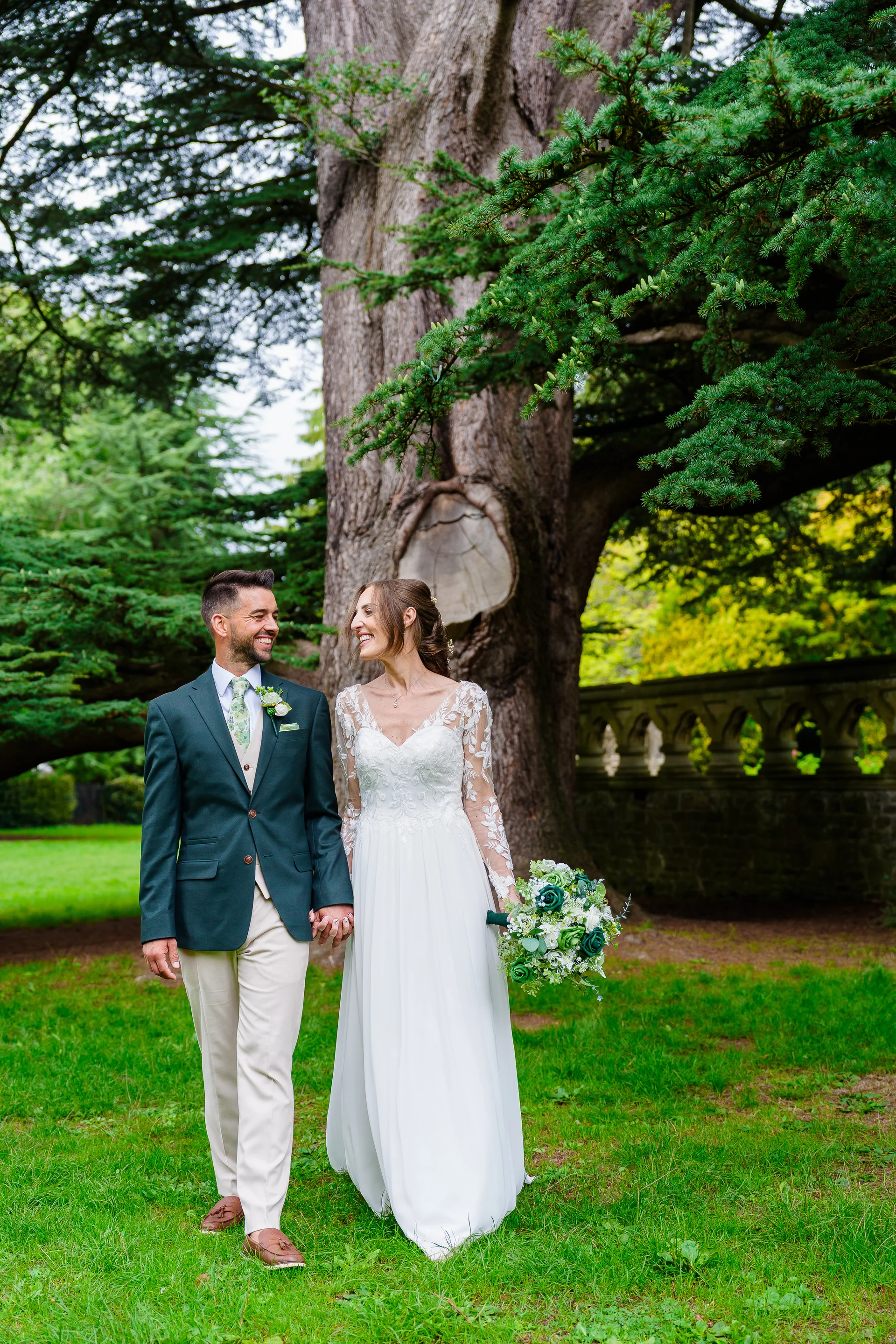 A smiling bride and groom walk hand in hand through lush green grounds beneath the branches of an ancient tree, with the bride holding a white and green bouquet.