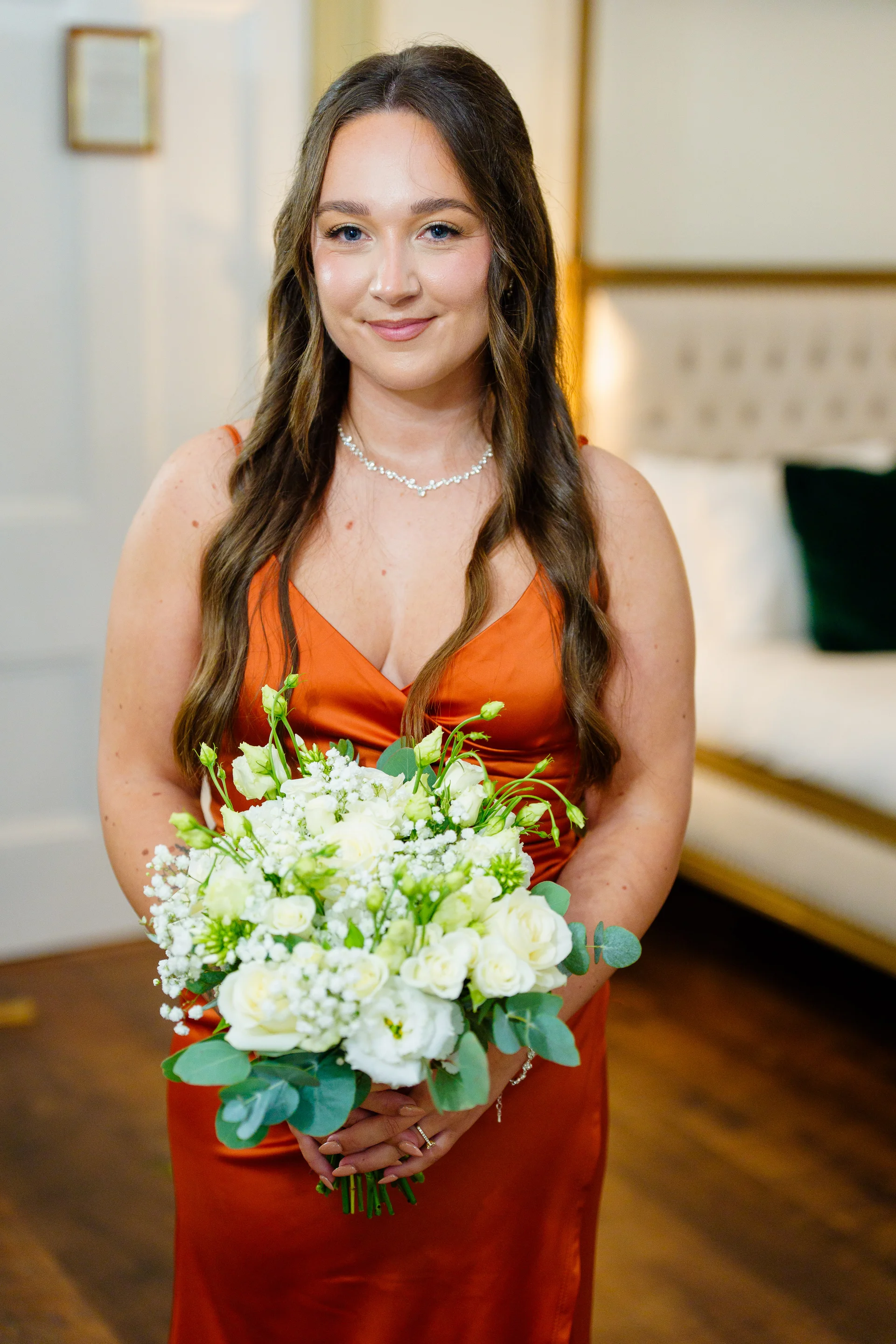 A smiling bridesmaid with long wavy hair holds a vibrant white floral bouquet, wearing an elegant rust-coloured satin dress.