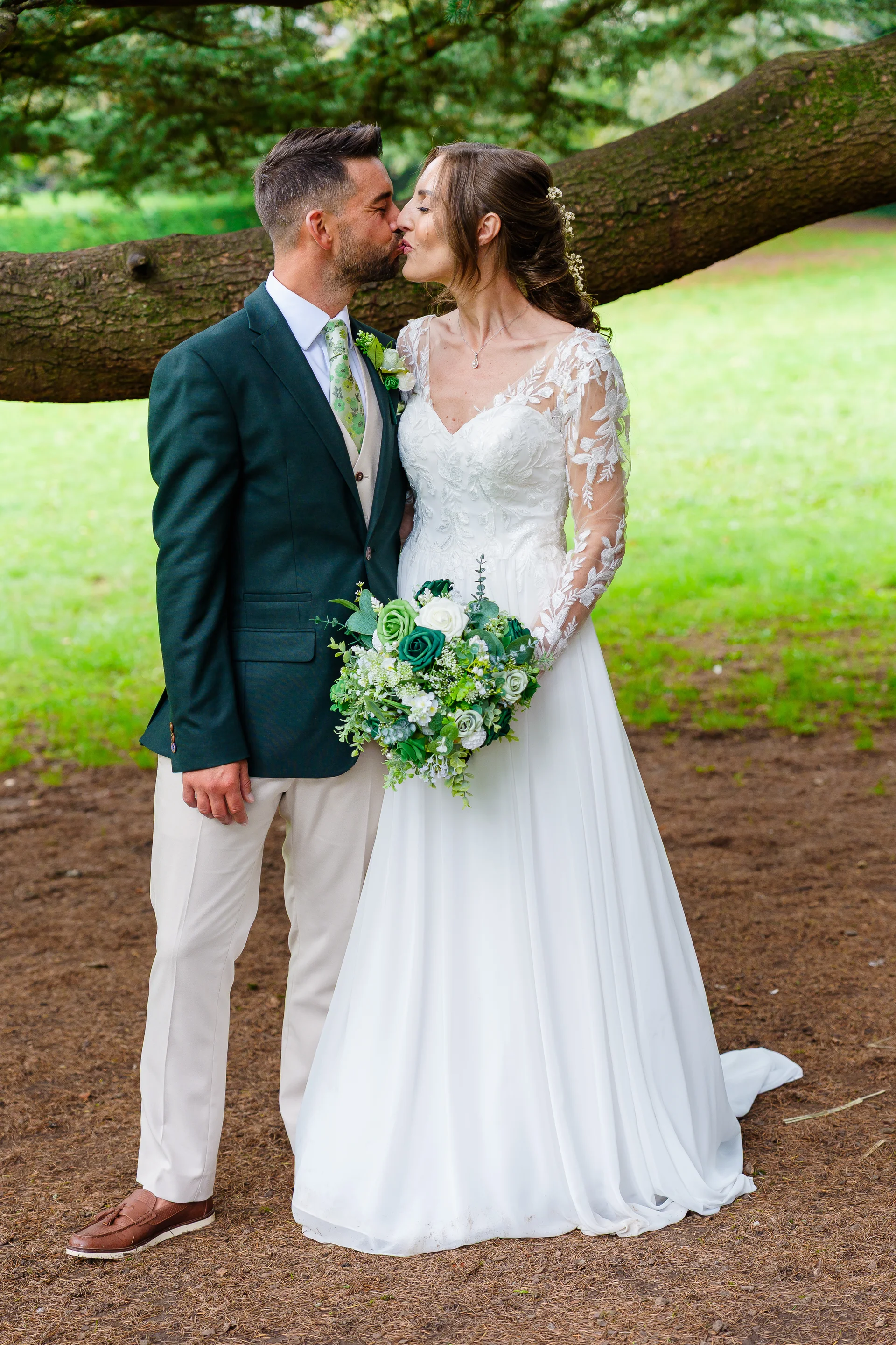 A bride and groom share a tender kiss beneath a large tree branch in a woodland setting, with the bride holding a lush green and white bouquet.
