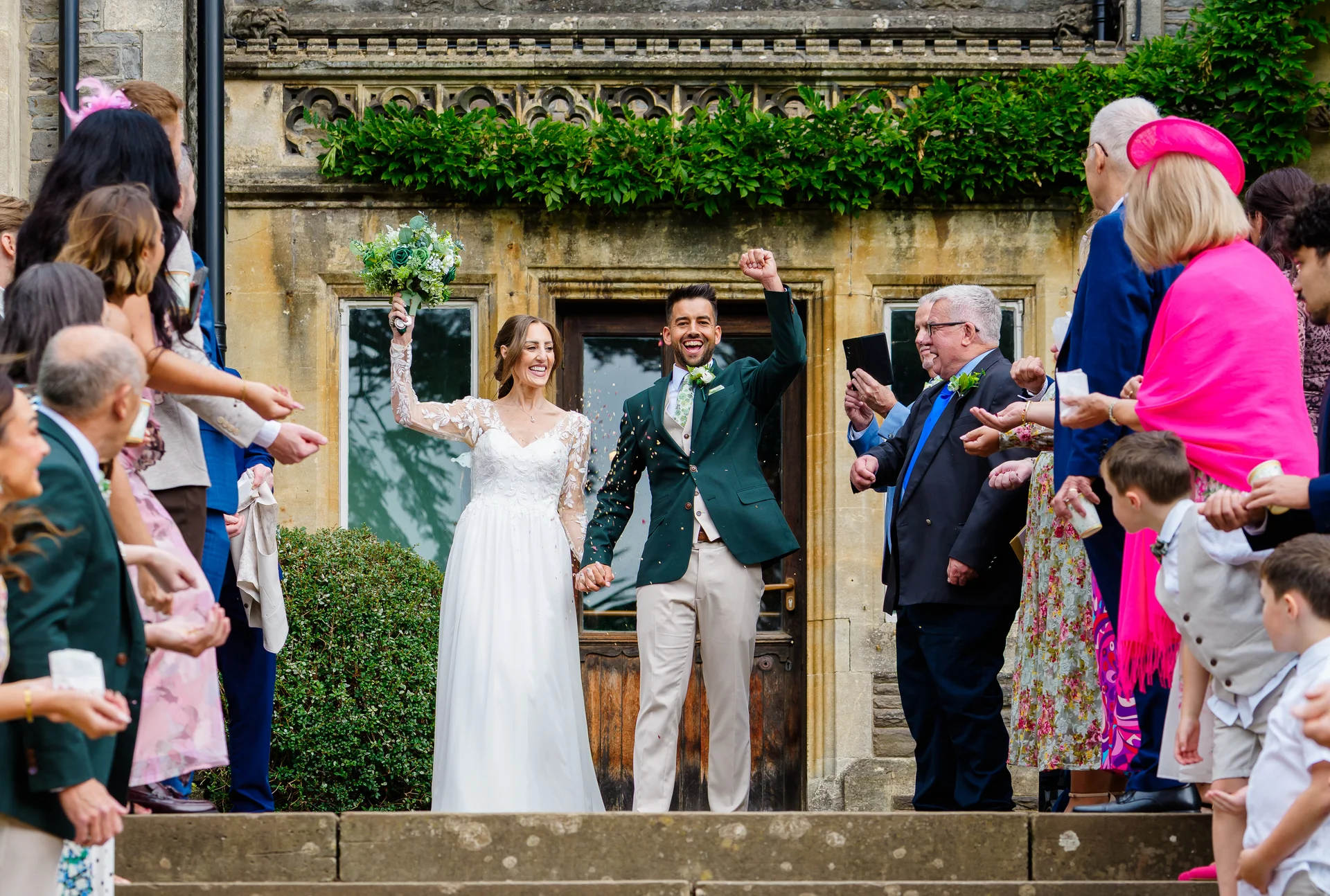A joyous newlywed couple exit a historic stone venue hand in hand while enthusiastic wedding guests shower them with celebratory confetti.