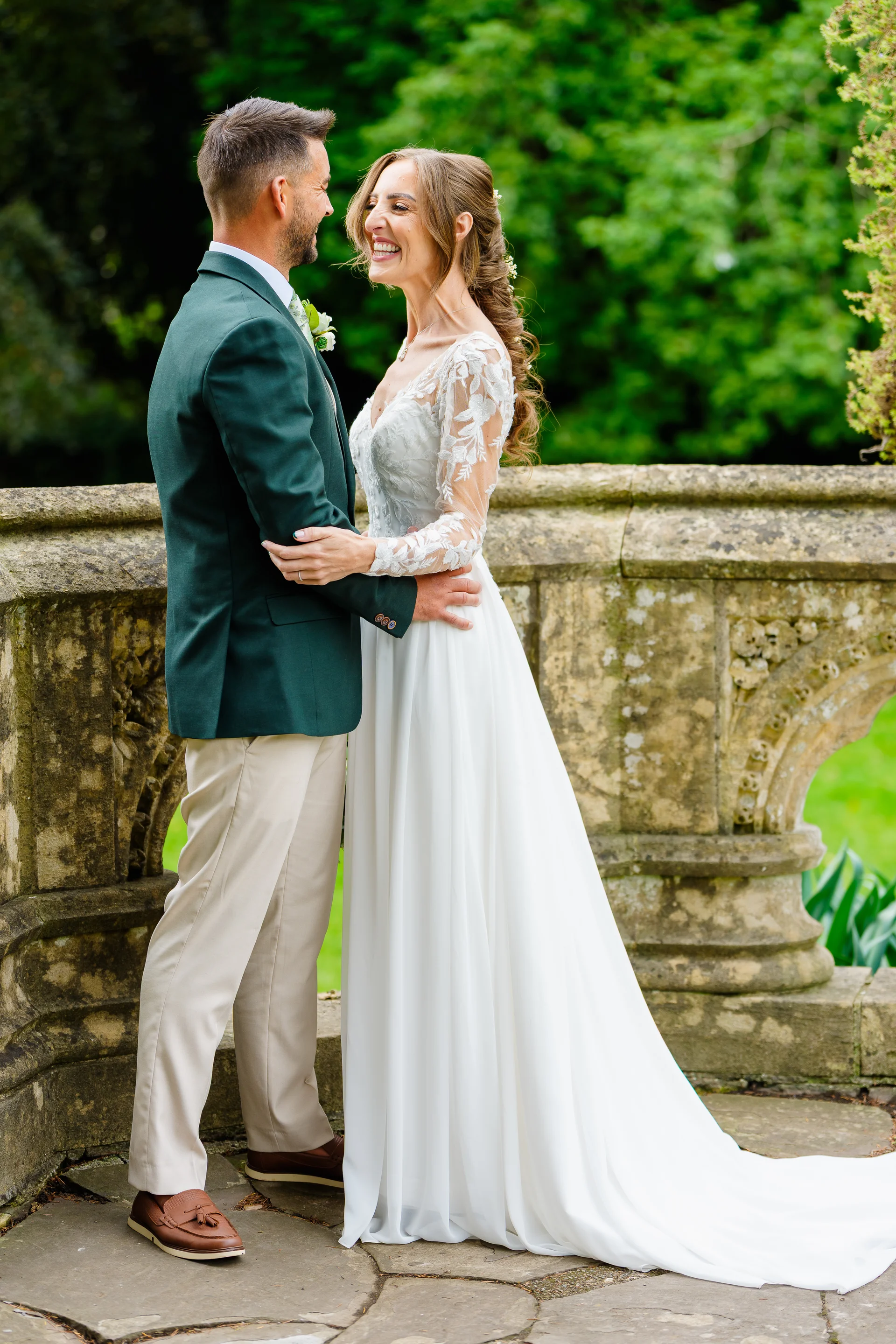 A joyful newlywed couple stand face-to-face laughing together outdoors on a stone terrace surrounded by lush greenery.
