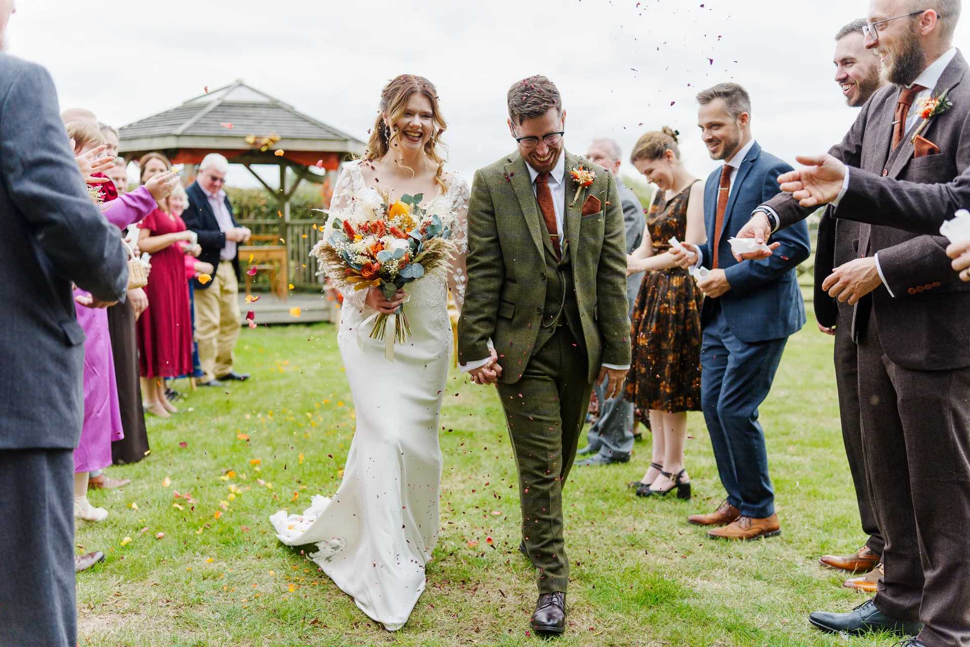 A joyful newlywed couple walks hand in hand through a shower of colourful confetti thrown by smiling guests outdoors on a lush green lawn.